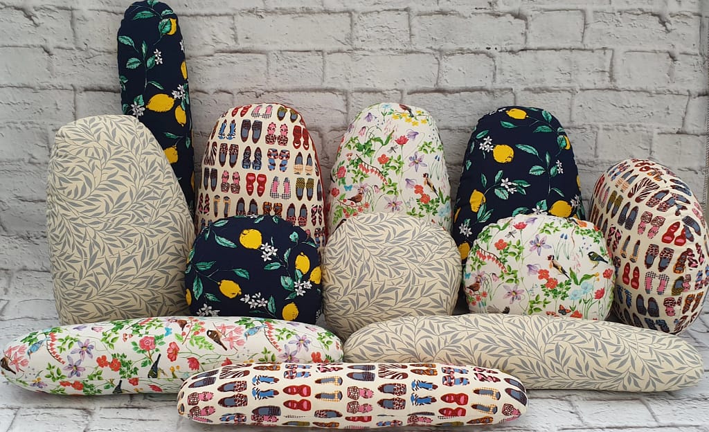 “Handmade tailor’s hams and sleeve rolls in colourful patterned fabrics arranged against a white brick background.”
